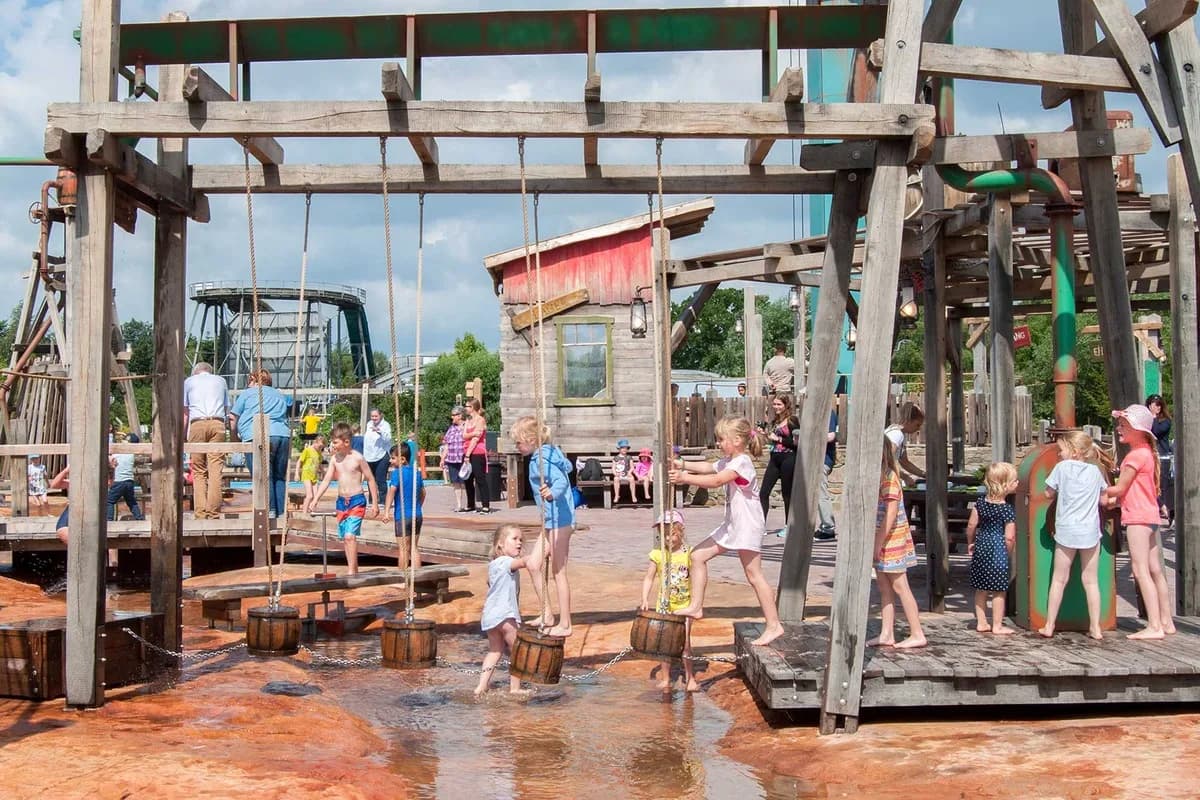 Wasserspielplatz „Yellowstone-Wackelpfad“ im Jaderpark — Kinder pumpen mit Holz-Eimern Wasser über einen wackeligen Holzsteg, Eltern stehen am Rand, im Hintergrund weitere Spielelemente und eine Achterbahn.